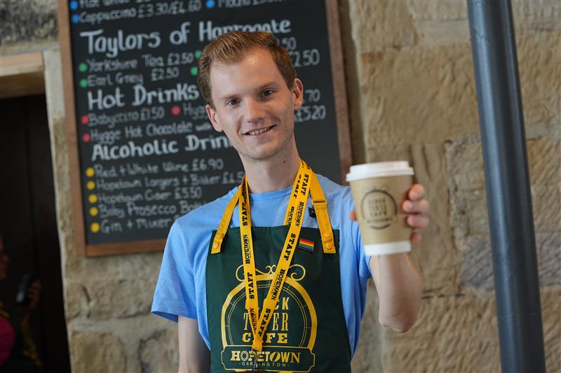 A member of staff Holding a takeout hot drink at the Hopetown Darlington Cafe. He's wearing a blue t-shirt, with a green apron and a yellow lanyard. He has short light brown hair.