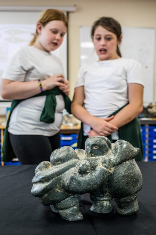 two girls in white t-shirt's talk together about the stone object of an Innuit family hugging in the table in front of them